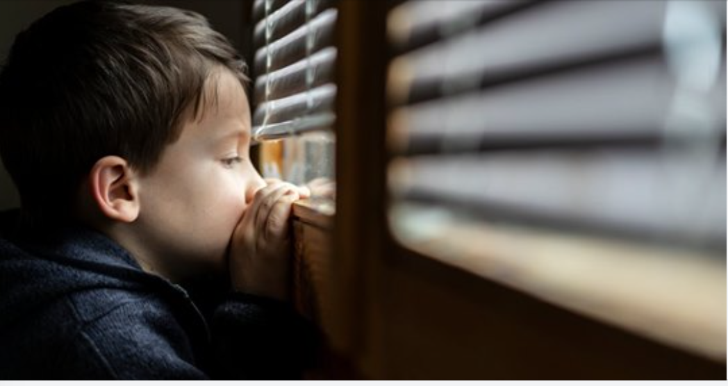 Young boy looking through the blinds of a window