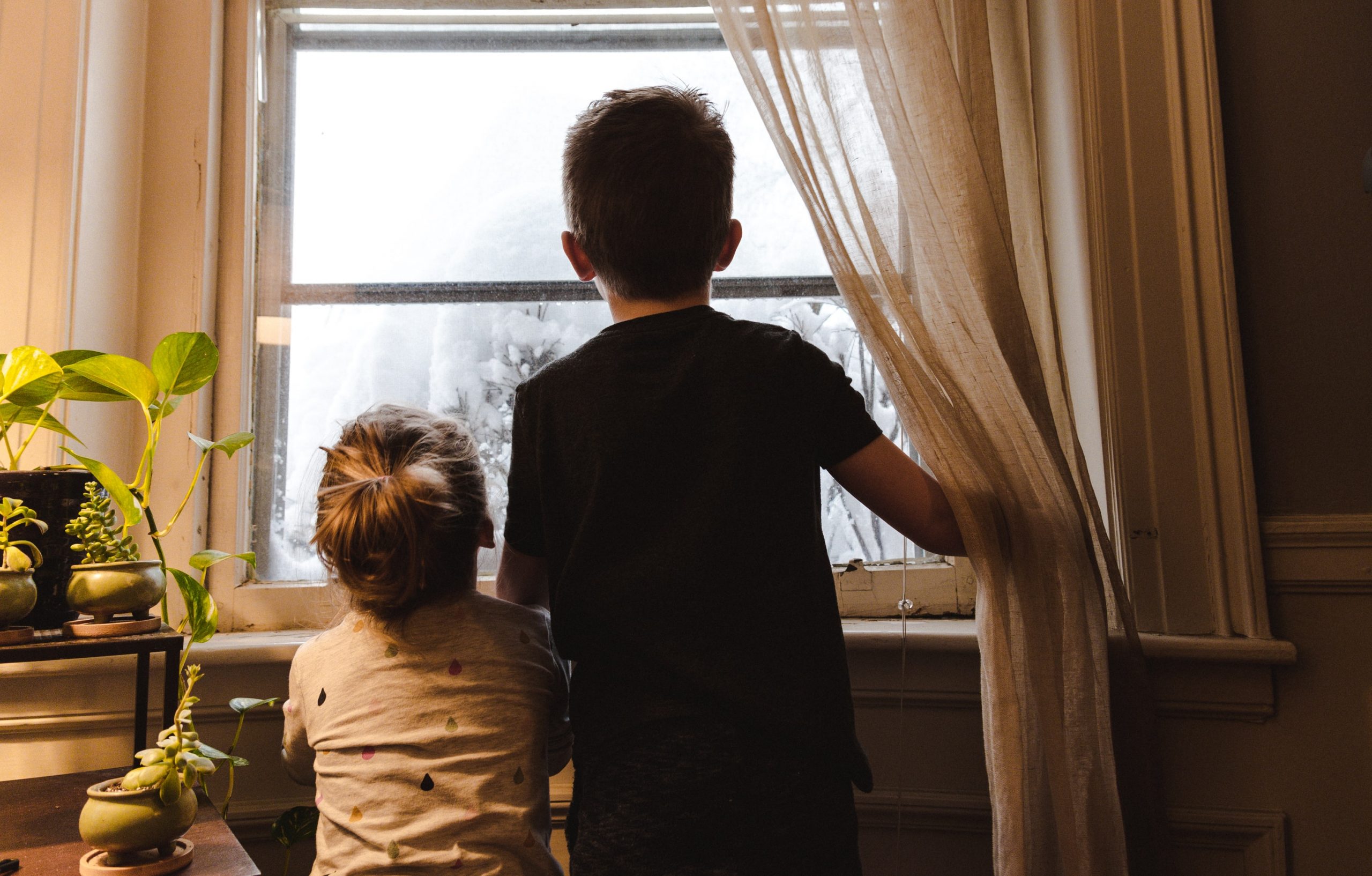 Two children hold a white curtain back and look out a window