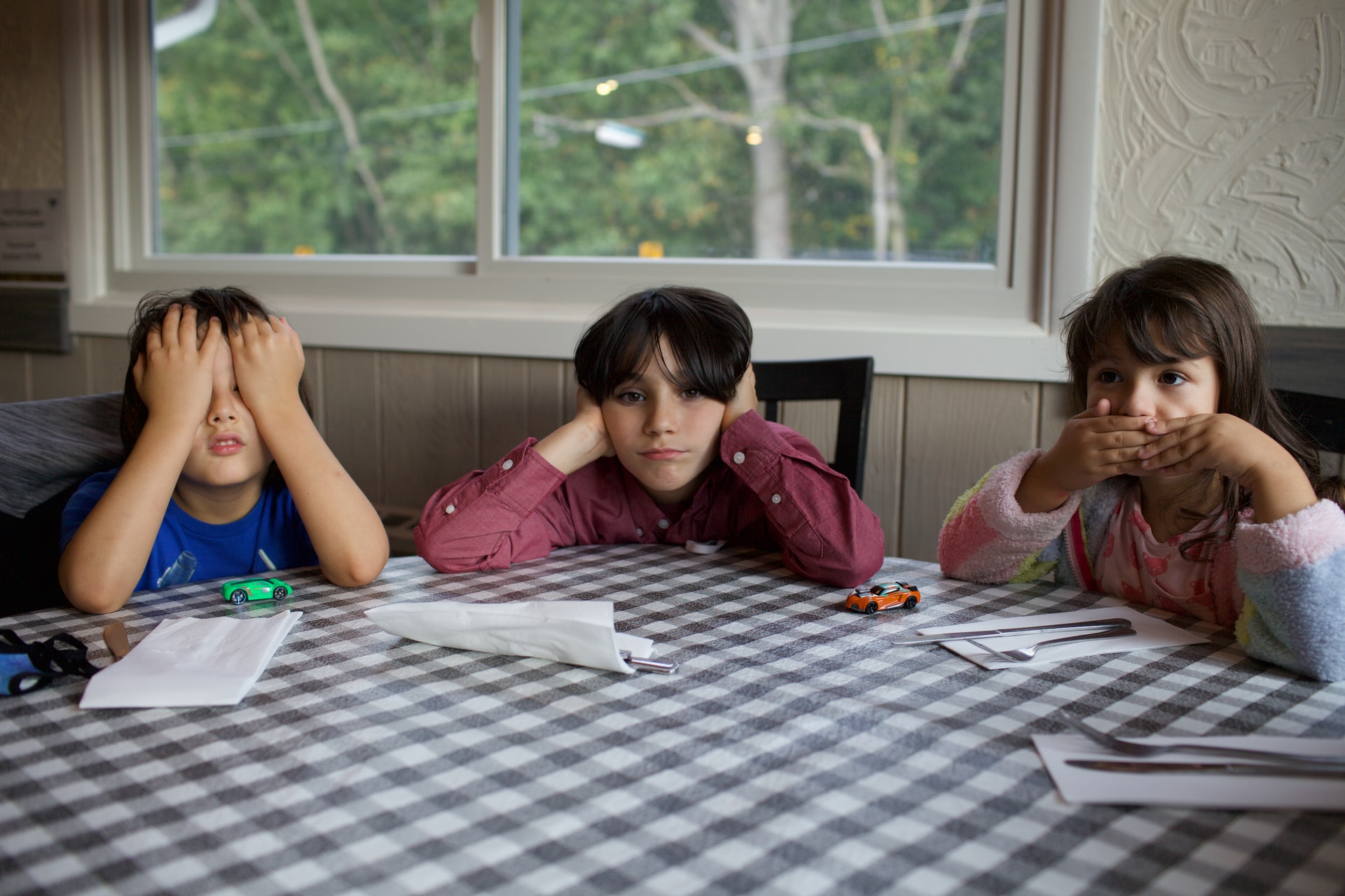 Thre kids sit at a table covering their eyes, ears, and nose