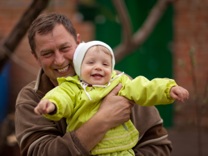 An older man smiling man holds a smiling baby dressed in yellow