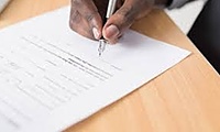 The fingers of a black male hold a pen of a paper to sign on a wooden desk